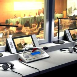 Pogled na konferencijsku salu iz prevodilačke kabine na čijem stolu su postavljeni prevodilački pultevi sa mikrofonima i slušalicama, te portabl video monitori i prevodilačke rasvetne lampe